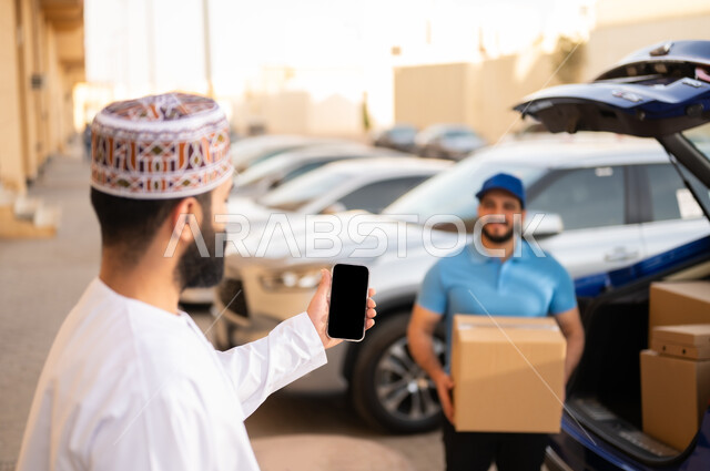 مفهوم التسوق الالكتروني, عملية توصيل الطلبات للزبائن، طلب المنتجات عبر الإنترنت, شاب عربي خليجي عماني يرتدي الدشداشة و الكمة يمسك بيده الهاتف المحمول, مندوب يحمل بيده صندوق مشتريات, عرض شاشة سوداء فارغة عبر الجوال