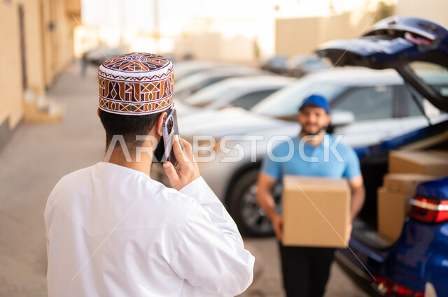 The process of delivering orders to customers, ordering products online, shipping services in Amman, an Arab Gulf Omani young man wearing a dishdasha and a kabbah making a phone call via mobile phone, a representative holding a shopping box in his hand