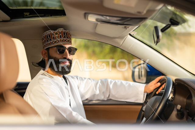 مفهوم القيادة الآمنة, النظر إلى الكاميرا بتعابير السعادة و السرور, الالتزام بوضع حزام الأمان, سائق عربي خليجي عماني يرتدي الدشداشة و الكمة مع نظارات شمسية يمسك المقود بيده و يقود السيارة بتعابير الفرح, خدمات النقل و التوصيل في عمان