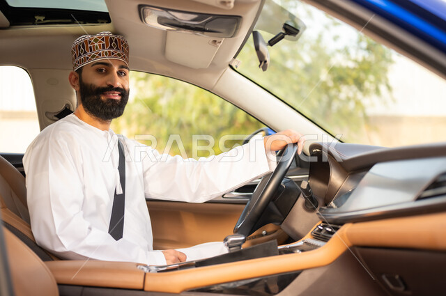 مفهوم القيادة الآمنة, النظر إلى الكاميرا بتعابير السعادة و السرور, الالتزام بوضع حزام الأمان, سائق عربي خليجي عماني يرتدي الدشداشة و الكمة يمسك المقود بيده و يقود السيارة بتعابير الفرح, خدمات النقل و التوصيل في عمان