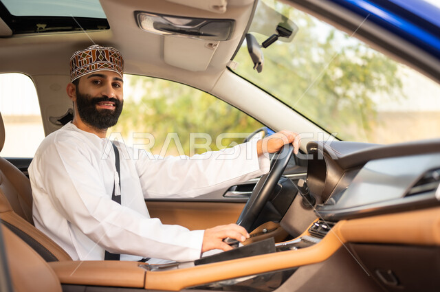 النظر إلى الكاميرا بتعابير السعادة و السرور, الالتزام بوضع حزام الأمان, مفهوم القيادة الآمنة, سائق عربي خليجي عماني يرتدي الدشداشة و الكمة يمسك المقود بيده و يقود السيارة بتعابير الفرح, خدمات النقل و التوصيل في عمان