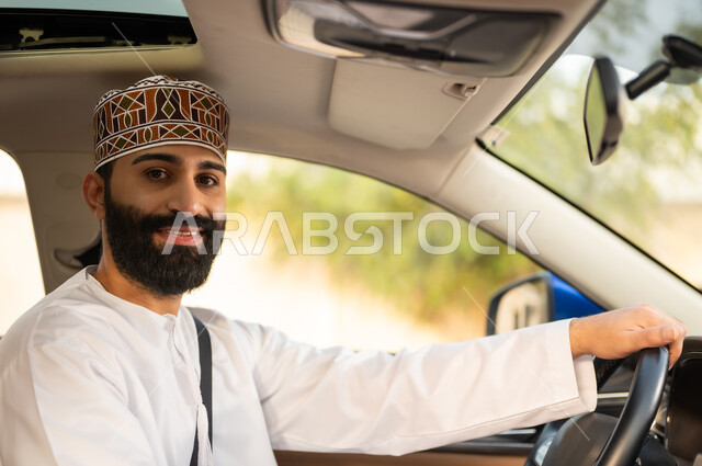 Looking at the camera with expressions of happiness and pleasure, commitment to wearing a seat belt, the concept of safe driving, a close-up of an Arab Gulf-Omani driver wearing a dishdasha and a sleeve, holding the steering wheel in his hand and driving the car with expressions of joy, transportation and delivery services in Amman