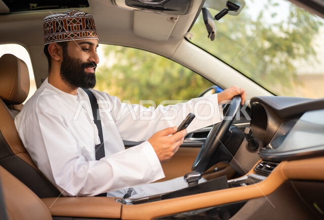 استخدام التقنيات الحديثة لطلب التاكسي من خلال تطبيق التوصيل، خدمات النقل الآمنة, سائق عربي خليجي عماني يرتدي الدشداشة و الكمة يقود السيارة و يستخدم الهاتف المحمول لتحديد موقع الركاب, مفهوم القيادة في عمان