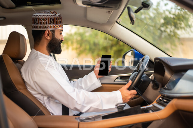 مفهوم القيادة في عمان, خدمات النقل الآمنة, استخدام التقنيات الحديثة لطلب التاكسي من خلال تطبيق التوصيل, سائق عربي خليجي عماني يرتدي الدشداشة و الكمة يجلس في السيارة و يمسك الهاتف المحمول بيده, عرض شاشة سوداء فارغة عبر الجوال