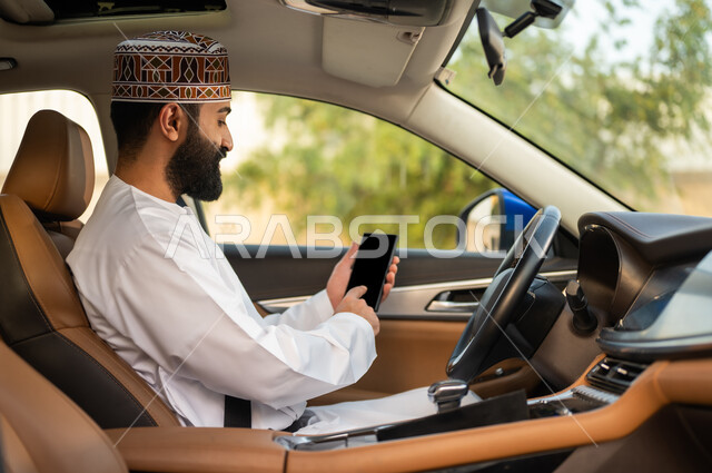 Safe transportation services, using modern technologies to request a taxi through the delivery application, an Arab Gulf Omani driver wearing a dishdasha and a kuma sitting in the car and holding a mobile phone in his hand, the concept of driving in Oman, displaying a blank black screen on the mobile phone