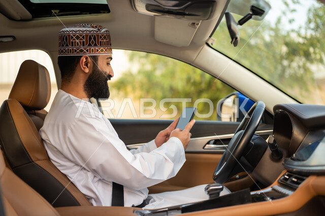 استخدام التقنيات الحديثة لطلب التاكسي من خلال تطبيق التوصيل، خدمات النقل الآمنة, سائق عربي خليجي عماني يرتدي الدشداشة و الكمة يقود السيارة و يستخدم الهاتف المحمول لتحديد موقع الركاب, مفهوم القيادة في عمان