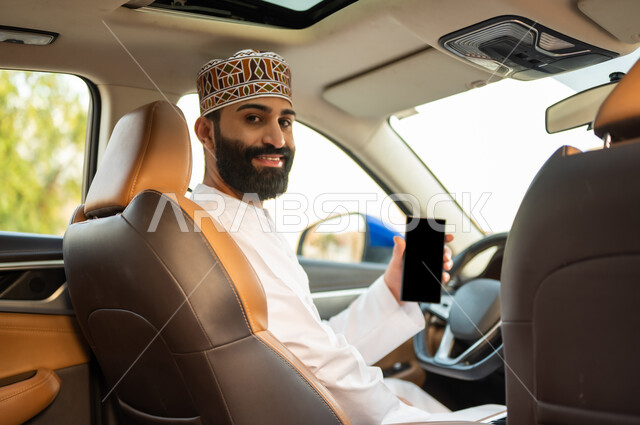 Using modern technologies to request a taxi through the delivery application, safe transportation services, an Arab Gulf Omani driver wearing a dishdasha and a kabbah sitting in the car and holding a mobile phone in his hand, the concept of driving in Oman, displaying a blank black screen on the mobile phone