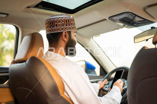 استخدام التقنيات الحديثة لطلب التاكسي من خلال تطبيق التوصيل، خدمات النقل الآمنة, سائق عربي خليجي عماني يرتدي الدشداشة و الكمة يقود السيارة و يستخدم الهاتف المحمول لتحديد موقع الركاب, مفهوم القيادة في عمان