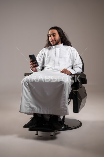 الدردشة مع العائلة و الأصدقاء عبر الجوال, تصفح مواقع التواصل الاجتماعي, استخدام الأجهزة التقنية الحديثة للتسلية و الترفيه, بورتريه لشاب عربي خليجي سعودي يرتدي الثوب التقليدي يجلس على الكرسي و يمسك الهاتف المحمول بيده, الجسم بالطول الكامل, خلفية رمادية