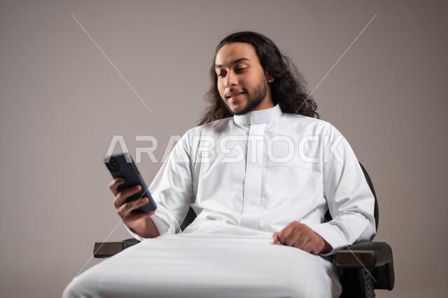 Chat with family and friends via mobile phone, browsing social networking sites, using modern technical devices for fun and entertainment, portrait of a Saudi Gulf Arab young man wearing a traditional dress sitting on a chair and holding a mobile phone in his hand, gray background