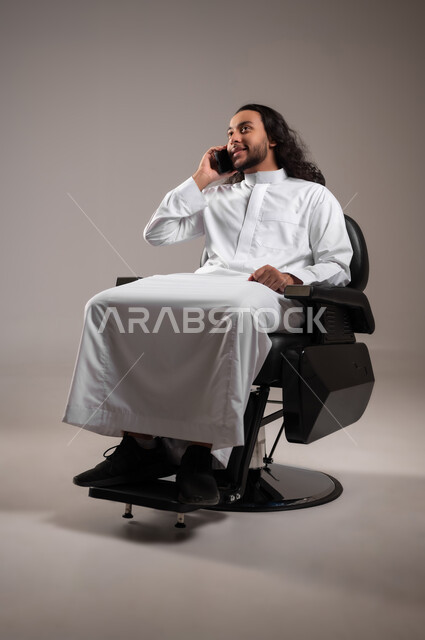إجراء مكالمة هاتفية عبر الجوال, تبادل الأحاديث و الأخبار الجيدة, التواصل مع العائلة و الأصدقاء, بورتريه لشاب عربي خليجي سعودي يرتدي الثوب التقليدي يجلس على الكرسي و يمسك الهاتف المحمول بيده, الجسم بالطول الكامل, خلفية رمادية
