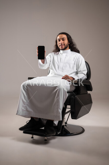 استخدام الأجهزة التقنية الحديثة, النظر إلى الكاميرا بإيماءات السعادة و السرور, بورتريه لشاب عربي خليجي سعودي مبتسم يرتدي الثوب التقليدي يجلس على الكرسي و يمسك الهاتف المحمول بيده, عرض شاشة سوداء فارغة عبر الجوال, الجسم بالطول الكامل, خلفية رمادية