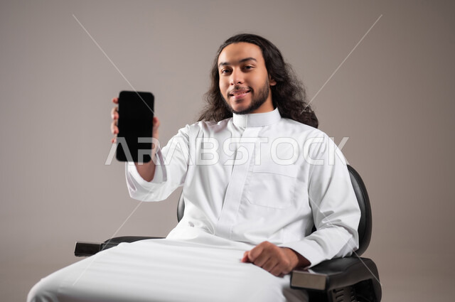 استخدام الأجهزة التقنية الحديثة للتسلية و الترفيه, النظر إلى الكاميرا بإيماءات السعادة و السرور, بورتريه لشاب عربي خليجي سعودي مبتسم يرتدي الثوب التقليدي يجلس على الكرسي و يمسك الهاتف المحمول بيده, عرض شاشة سوداء فارغة عبر الجوال, خلفية رمادية