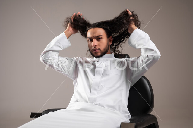 مفهوم الاهتمام و العناية بالنظافة الشخصية, الحاجة للذهاب إلى الحلاق لتغيير المظهر الخارجي, بورتريه مقرب لشاب عربي خليجي سعودي يرتدي الثوب التقليدي ينظر إلى الكاميرا بتعابير الجدية و يمسك بيديه شعره الطويل, خلفية رمادية