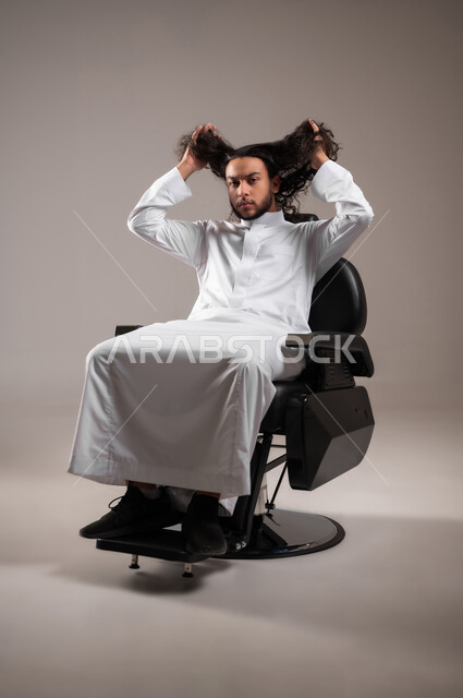 The concept of attention and care for personal hygiene, the need to go to the barber to change the external appearance, a portrait of a Saudi Gulf Arab young man wearing the traditional dress, looking at the camera with a serious expression and holding his long hair in his hands, full-length body, gray background