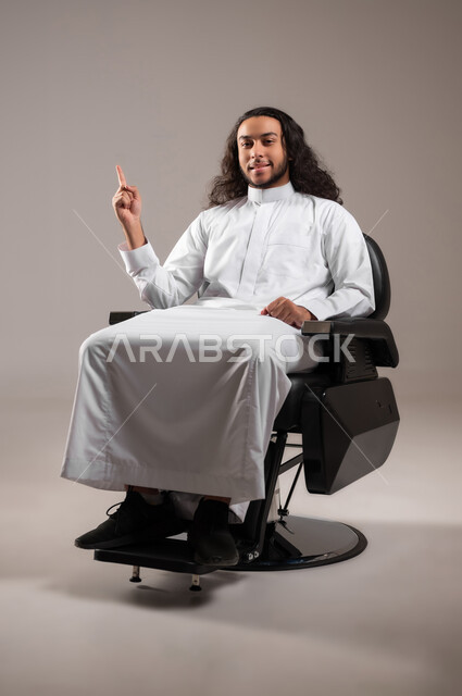 إشارة تمثل الرقم واحد, النظر إلى الكاميرا بإيماءات السعادة و السرور, بورتريه لشاب عربي خليجي سعودي مبتسم يرتدي الثوب التقليدي يجلس على الكرسي و يشير بإصبع السبابة للأعلى, الجسم بالطول الكامل, خلفية رمادية