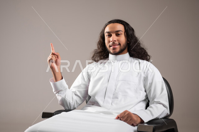 إشارة تمثل الرقم واحد, النظر إلى الكاميرا بإيماءات السعادة و السرور, بورتريه مقرب لشاب عربي خليجي سعودي مبتسم يرتدي الثوب التقليدي يجلس على الكرسي و يشير بإصبع السبابة للأعلى, خلفية رمادية