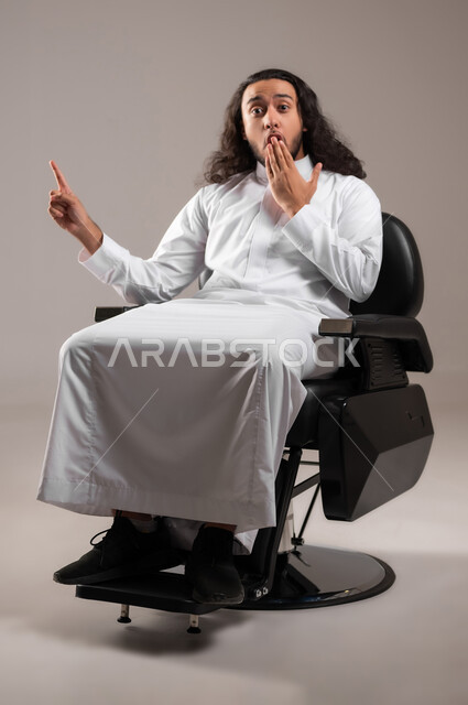 النظر إلى الكاميرا بإيماءات الدهشة و الذهول, إشارة تمثل الرقم واحد, بورتريه لشاب عربي خليجي سعودي يرتدي الثوب التقليدي يجلس على الكرسي و يشير بإصبع السبابة للأعلى, الجسم بالطول الكامل, خلفية رمادية
