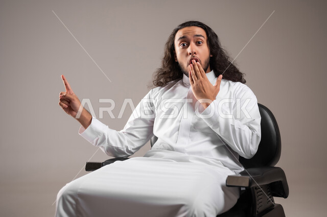 النظر إلى الكاميرا بإيماءات الدهشة و الذهول, إشارة تمثل الرقم واحد, بورتريه مقرب لشاب عربي خليجي سعودي يرتدي الثوب التقليدي يجلس على الكرسي و يشير بإصبع السبابة للأعلى, خلفية رمادية