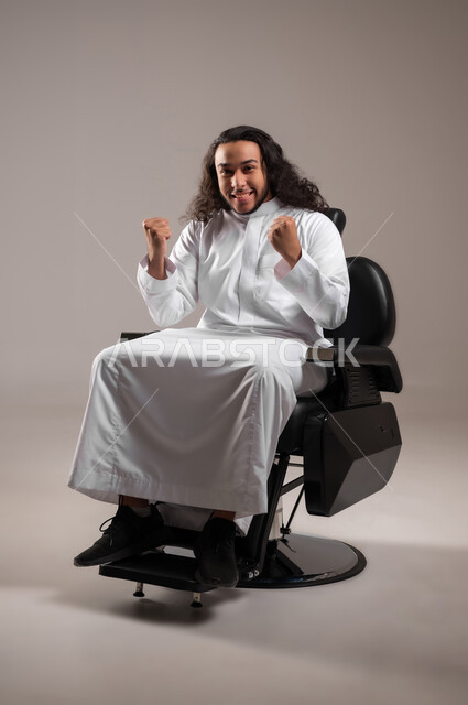 إيماءات الفرح لسماع خبر ما, النظر الى الكاميرا بتعابير الفوز و تحقيق الأهداف, بورتريه لشاب عربي خليجي سعودي يرتدي الثوب التقليدي يجلس على الكرسي و يرفع يديه بحركات الحماس و السعادة,, الجسم بالطول الكامل, خلفية رمادية