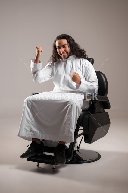 إيماءات الفرح لسماع خبر ما, النظر الى الكاميرا بتعابير الفوز و تحقيق الأهداف, بورتريه لشاب عربي خليجي سعودي يرتدي الثوب التقليدي يجلس على الكرسي و يرفع يديه بحركات الحماس و السعادة, الجسم بالطول الكامل, خلفية رمادية