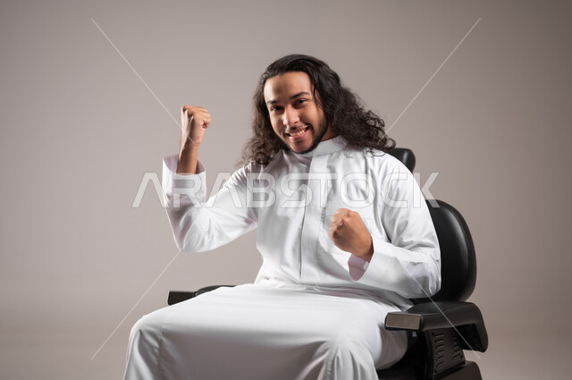 Looking at the camera with expressions of winning and achieving goals, gestures of joy at hearing some news, portrait of a Saudi Gulf Arab young man wearing a traditional dress sitting on a chair and raising his hands with movements of enthusiasm and happiness, gray background