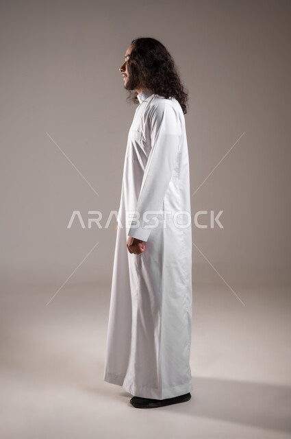 مفهوم العناية بالنظافة الشخصية, الاهتمام بالمظهر الخارجي, بورتريه من الجانب لشاب عربي خليجي سعودي يرتدي الثوب التقليدي يقف باستقامة و اعتدال, الجسم بالطول الكامل, خلفية رمادية