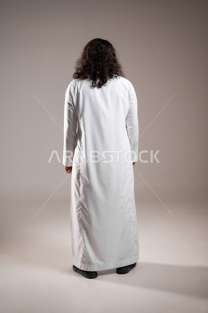 الاهتمام بالمظهر الخارجي, مفهوم العناية بالنظافة الشخصية, بورتريه من الخلف لشاب عربي خليجي سعودي يرتدي الثوب التقليدي يقف باستقامة و اعتدال, الجسم بالطول الكامل, خلفية رمادية