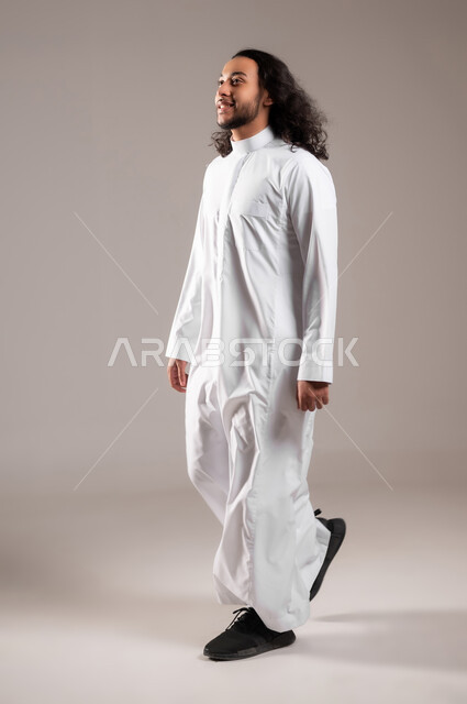 Going to the barber to change the external appearance, the concept of attention and care for personal hygiene, portrait of a smiling Saudi Arabian Gulf man wearing a traditional dress walking to the barbershop, full-length body, gray background