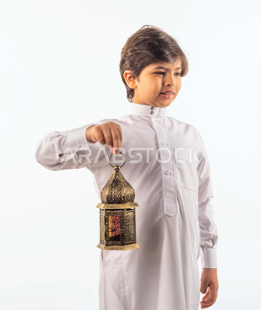 بورتريه لصبي صغير سعودي خليجي جميل ، يحمل بيده فانوس ملون بإيماءات وجه ويدين مختلفة , منظر جانبي لطفل سعودي خليجي يرتدي الزي السعودي التقليدي الأنيق , العبادة والتقرب إلى الله , فانوس رمضان , خلفية بيضاء