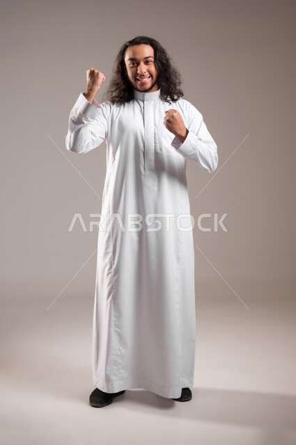 النظر الى الكاميرا بتعابير الفوز و تحقيق الأهداف، إيماءات الفرح لسماع خبر ما, بورتريه لشاب عربي خليجي سعودي يرتدي الثوب التقليدي يرفع يديه بحركات الحماس و السعادة, الجسم بالطول الكامل, خلفية رمادية