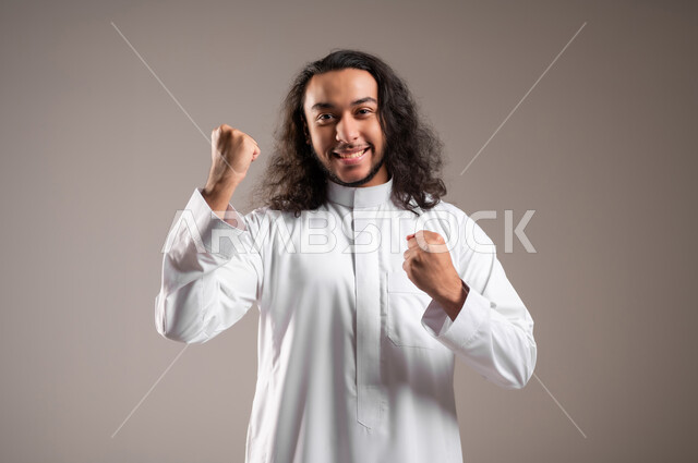 النظر الى الكاميرا بتعابير الفوز و تحقيق الأهداف، إيماءات الفرح لسماع خبر ما, بورتريه مقرب لشاب عربي خليجي سعودي يرتدي الثوب التقليدي يرفع يديه بحركات الحماس و السعادة, خلفية رمادية