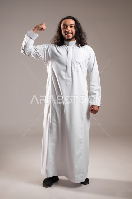 ممارسة الرياضة للحصول على جسم سليم, استعراض عضلات اليدين, مفهوم اللياقة البدنية, بورتريه لشاب عربي خليجي سعودي يرتدي الثوب التقليدي يقف باستقامة بحركات تدل على القوه و الصحة الجسدية, الجسم بالطول الكامل, خلفية رمادية