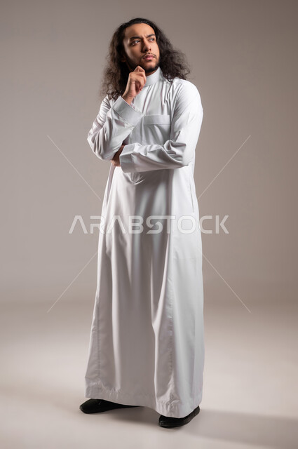 التخطيط للمستقبل وشرود الذهن، محاولة إيجاد حل لمشكلة ما،, بورتريه لشاب عربي خليجي سعودي يرتدي الثوب التقليدي يضع يده تحت ذقنه بإيماءات التفكير و التركيز, الجسم بالطول الكامل, خلفية رمادية