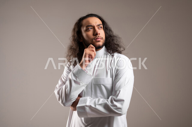 التخطيط للمستقبل وشرود الذهن، محاولة إيجاد حل لمشكلة ما،, بورتريه مقرب لشاب عربي خليجي سعودي يرتدي الثوب التقليدي يضع يده تحت ذقنه بإيماءات التفكير و التركيز, خلفية رمادية