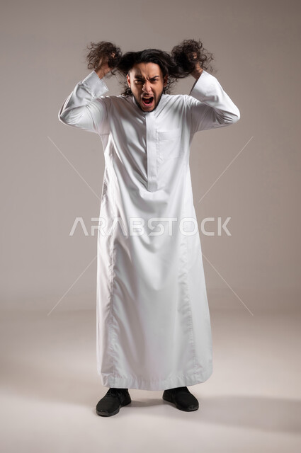 الحاجة للذهاب إلى الحلاق لتغيير المظهر الخارجي, إيماءات الغضب و التوتر و الضغط النفسي, بورتريه لشاب عربي خليجي سعودي يرتدي الثوب التقليدي يمسك بيديه شعره الطويل بتعابير الصراخ الشديد, مفهوم الاهتمام و العناية بالنظافة الشخصية, الجسم بالطول الكامل, خلفية رمادية
