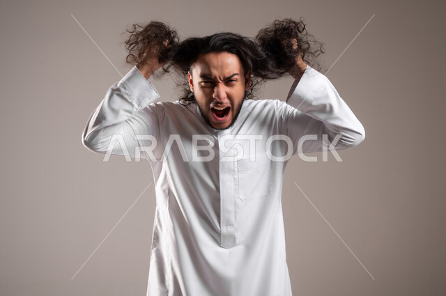 الحاجة للذهاب إلى الحلاق لتغيير المظهر الخارجي, إيماءات الغضب و التوتر و الضغط النفسي, بورتريه مقرب لشاب عربي خليجي سعودي يرتدي الثوب التقليدي يمسك بيديه شعره الطويل بتعابير الصراخ الشديد, مفهوم الاهتمام و العناية بالنظافة الشخصية, خلفية رمادية