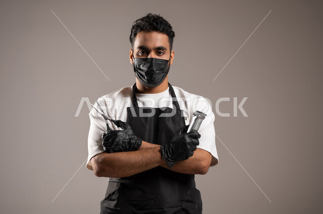 استخدام أدوات و معدات الحلاقة, صالون تجميلي رجالي لتصفيف الشعر, تقديم خدمات العناية الشخصية, بورتريه مقرب لحلاق عربي خليجي سعودي يرتدي زي الحلاقين التقليدي مع الكمامة يمسك بيديه المقص و الماكينة, خلفية رمادية