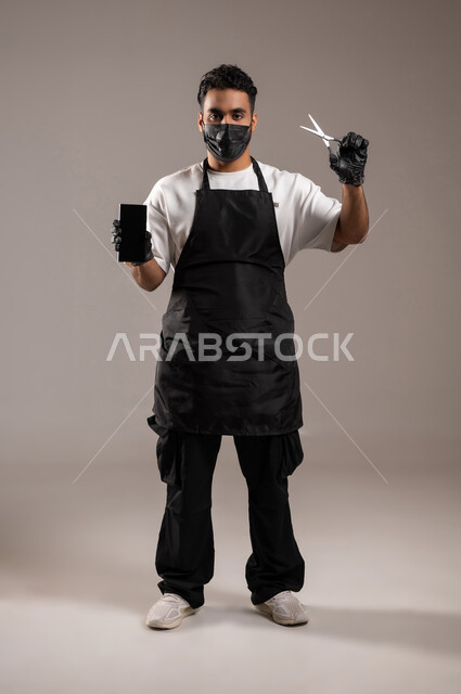 استخدام الأجهزة التقنية الحديثة, صالون حلاقة تجميلي رجالي, بورتريه لحلاق عربي خليجي سعودي يرتدي زي الحلاقين التقليدي مع الكمامة يمسك بيده المقص و الهاتف المحمول, عرض شاشة سوداء فارغة عبر الجوال, الجسم بالطول الكامل, خلفية رمادية