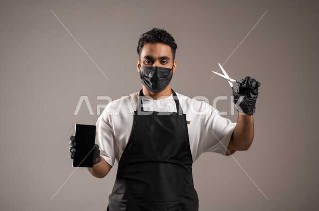 استخدام الأجهزة التقنية الحديثة, صالون حلاقة تجميلي رجالي، العناية و الاهتمام بشعر صحي ونظيف, بورتريه مقرب لحلاق عربي خليجي سعودي يرتدي زي الحلاقين التقليدي مع الكمامة يمسك بيده المقص و الهاتف المحمول, عرض شاشة سوداء فارغة عبر الجوال, خلفية رمادية