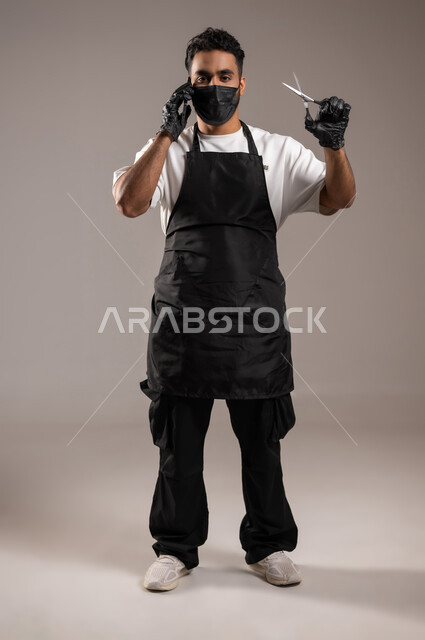 تقديم خدمات العناية الشخصية, صالون حلاقة لقص و تصفيف الشعر للرجال, مهن و وظائف في المملكة العربية السعودية, بورتريه مقرب لحلاق عربي خليجي يرتدي زي الحلاقين التقليدي مع الكمامة ينظر إلى الكاميرا و يمسك بيده المقص, الجسم بالطول الكامل, خلفية رمادية