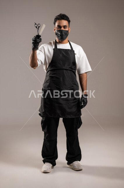 Providing personal care services, a barber shop for cutting and styling hair for men, professions and jobs in the Kingdom of Saudi Arabia, a close-up portrait of a Gulf Arab barber wearing a traditional barber uniform with a mask, looking at the camera and holding scissors and a comb in his hand, full-length body, gray background
