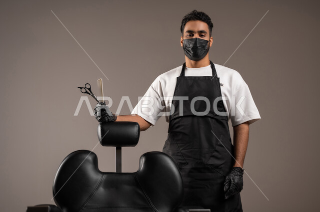 تقديم خدمات العناية الشخصية, مهن و وظائف في المملكة العربية السعودية, ممارسة مهنة الحلاقة, بورتريه لحلاق عربي خليجي سعودي يرتدي زي الحلاقين التقليدي مع الكمامة ينظر إلى الكاميرا و يمسك المقص و المشط بيده, خلفية رمادية