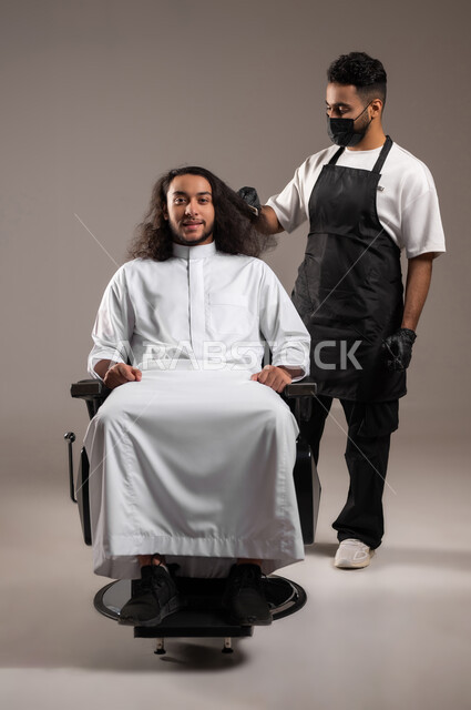 ممارسة مهنة الحلاقة, تقديم خدمات العناية الشخصية, الاهتمام بالأناقة و المظهر الخارجي, بورتريه لحلاق عربي خليجي يرتدي زي الحلاقين التقليدي مع القفازات يمشط شعر شاب سعودي باستخدام المشط, الجسم بالطول الكامل, خلفية رمادية
