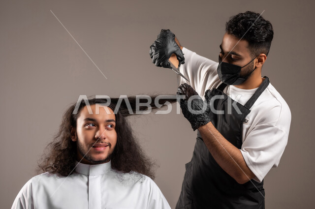 المحافظة على الأناقة والنظافة الشخصية، تقديم خدمات الحلاقة و تصفيف الشعر, الاهتمام بالمظهر الخارجي, بورتريه مقرب لحلاق عربي خليجي يرتدي زي الحلاقين التقليدي مع الكمامة يحلق لشاب سعودي باستخدام المقص, خلفية رمادية