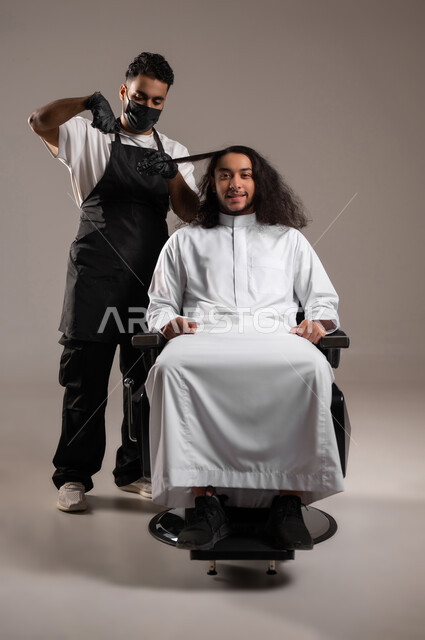 المحافظة على الأناقة والنظافة الشخصية، تقديم خدمات الحلاقة و تصفيف الشعر, الاهتمام بالمظهر الخارجي, بورتريه لحلاق عربي خليجي يرتدي زي الحلاقين التقليدي مع الكمامة يحلق لشاب سعودي باستخدام المقص, الجسم بالطول الكامل, خلفية رمادية