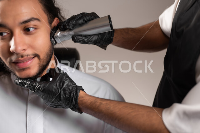تقديم خدمات العناية الشخصية, الاهتمام بالشعر و النظافة البدنية, ممارسة مهنة الحلاقة, بورتريه مقرب لحلاق عربي خليجي يرتدي زي الحلاقين التقليدي مع القفازات يحلق ذقن شاب سعودي باستخدام ماكينة الحلاقة, خلفية رمادية