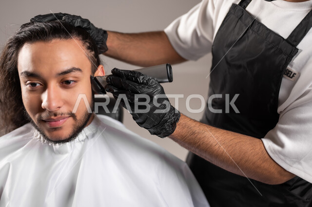 استعمال ماكينة الحلاقة, تقديم خدمات العناية الشخصية, الاهتمام بالشعر و النظافة البدنية, بورتريه مقرب لحلاق عربي خليجي يرتدي زي الحلاقين التقليدي مع القفازات يحلق لشاب سعودي, استخدام الغطاء الأبيض لحماية الملابس, خلفية رمادية