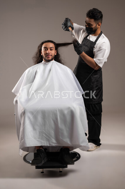 ممارسة مهنة الحلاقة, تقديم خدمات العناية الشخصية, الاهتمام بالشعر و النظافة البدنية, بروتريه لحلاق عربي خليجي يرتدي زي الحلاقين التقليدي مع الكمامة يحلق لشاب سعودي باستخدام المقص, استعمال الغطاء الأبيض لحماية الملابس, الجسم بالطول الكامل, خلفية رمادية