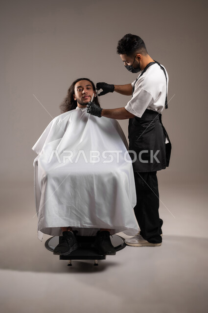استعمال المقص و المشط لحلاقة الذقن, تقديم خدمات العناية الشخصية, الاهتمام بالشعر و النظافة البدنية, بروتريه لحلاق عربي خليجي يرتدي زي الحلاقين التقليدي مع الكمامة يحلق لشاب سعودي, استخدام الغطاء الأبيض لحماية الملابس, الجسم بالطول الكامل, خلفية رمادية